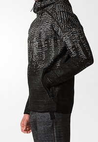 Haut à capuche noir en tissu texturé, avec une fermeture éclair sur le devant et des poches latérales. Le design comprend des motifs contrastés et une coupe ajustée.