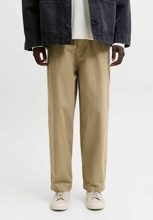 Personne portant un pantalon kaki à jambes larges, des baskets beige, un pull blanc et une veste en jean noire, debout sur un fond uni.