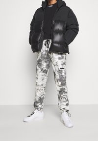 Svart vadderad jacka med huva, tillsammans med ljusgrå cargo-byxor med splattermönster och sidofickor, avslutat med vita sneakers.