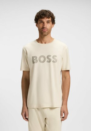 Mand med krøllet hår iført en lys beige kortærmet skjorte og bukser, skjorten har stor 'BOSS' tekst trykt på brystet.