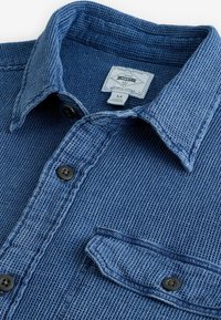 Cămașă texturată albastră cu guler cu nasturi, nasturi negri și un buzunar pe piept. Eticheta este vizibilă lângă guler. Design dintr-un material rezistent.