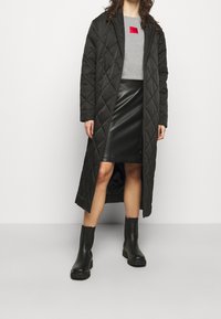 Žena v dlouhém černém prošívaném kabátu, šedém tričku s červeným logem, černé kožené sukni a černých robustních kotníkových botách na jednolitém pozadí.