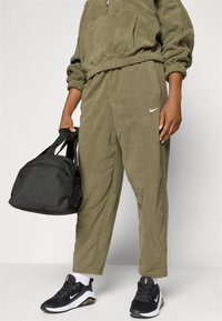 Olijfgroen fleece trainingspak met elastische manchetten, kangoeroezak en Nike-logo. Zwarte sporttas met schouderbanden. Zwart-witte sneakers.