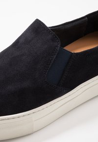Sapatilha slip-on em camurça azul marinho escura com painéis laterais elásticos, sola de borracha branca e detalhes de costura visíveis. Textura suave e design limpo.