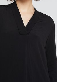Blusa negra con escote en V, de textura suave, mangas largas y un detalle sutil en los puños de las mangas.