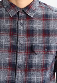 Camisa de cuadros en gris, azul marino y burdeos, con cuello abotonado, mangas largas y un bolsillo en el pecho que presenta un sutil logo.