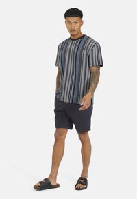 Koszulka w paski w czarnym, niebieskim i szarym kolorze z krótkimi rękawami, w połączeniu z ciemnymi szortami i czarnymi sandałami, stojąca na białym tle.