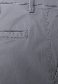 Harmaat kangast lyhyet housut, joissa on sileä pinta. Ominaisuuksiin kuuluu sivutasku läpällä, tikkausnuotit ja suora leikkaus.