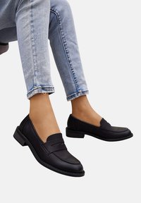 Mocassins en cuir noir avec un petit talon bloc, dotés de coutures sur le devant et d'une bande sur le cou-de-pied. Portés avec un jean en denim bleu clair.