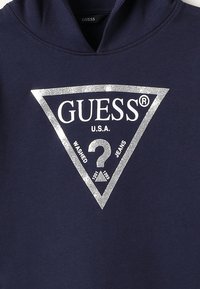 Sudadera con capucha azul marino con un gran logo de triángulo de purpurina plateada que dice "GUESS U.S.A." y texto "WASHED JEANS" en blanco. Textura de tela suave.