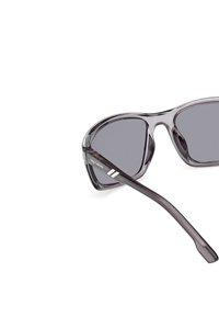 Gafas de sol con un marco gris transparente, lentes triangulares y lentes oscuros. En el lateral cuentan con la marca "SKECHERS" y acentos plateados.