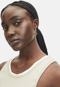 Goldene Creolen mit einem quadratischen Anhänger, zarte goldene Halskette mit einem quadratischen Charme und ein hellbeiges Tanktop mit weißen Akzenten.