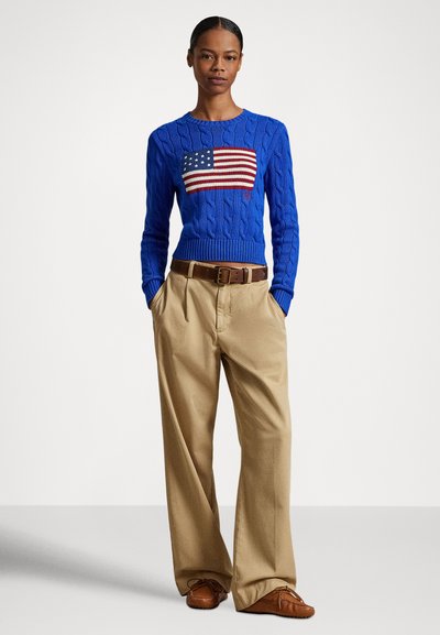 Kék kötött pulóver amerikai zászló mintával elöl; bézs, redőzött nadrággal és barna loaferekkel párosítva.