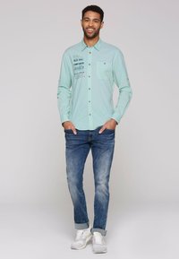 Camicia button-up azzurro chiaro realizzata in tessuto testurizzato, con dettagli stampati, una tasca sul petto sinistro, abbinata a jeans blu e sneakers bianche.