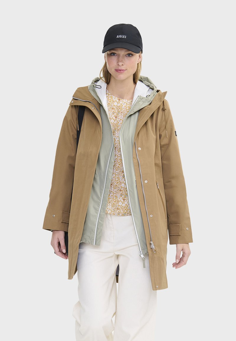 Brązowa wodoodporna kurtka z odpinanym jasnooliwkowym wnętrzem, zestawiona z kwiatowym topem. Model nosi czapkę w kolorze czarnym i off-white spodnie.
