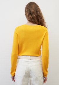 Femme aux longs cheveux bouclés portant un pull jaune vif et un pantalon blanc taille haute, debout contre un mur blanc uni, de dos au spectateur.