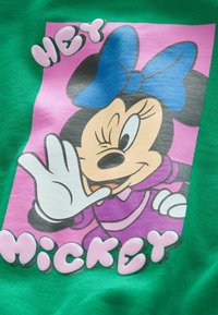Grøn T-shirt med et grafisk motiv af en tegneseriefigur med en blå sløjfe og lilla kjole, samt pink tekst i boblebogstaver, der står "HEY MICKEY."