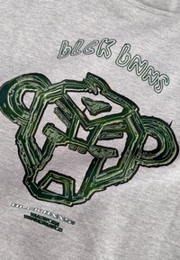 Grijs T-shirt met korte mouwen met een groot groen grafisch ontwerp van een gestileerd gezicht en vetgedrukte tekst "BLCK BNNS" in een speelse lettertype.