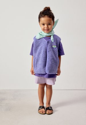 Jaunas vaikas stovi, dėvi violetinę trumpomis rankovėmis marškinėlį, levandų spalvos fliiso liemenę, šviesiai žalią šaliką, šortus ir juodus sandalus su sagtimis.