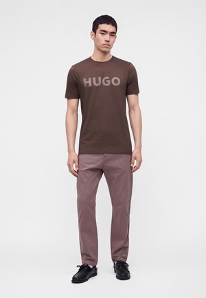 Млад мъж, облечен в кафява тениска HUGO, люлякови панталони и черни обувки, стои пред обикновен бял фон.