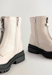 Bottines beige clair avec des semelles en caoutchouc noires épaisses et des tirettes en forme d'étoile sur un fond uni clair.