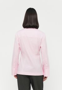 Lys rosa bluse med lange ermer, med rett snitt, knappdetalj på ermene, og et rent bakdesign.