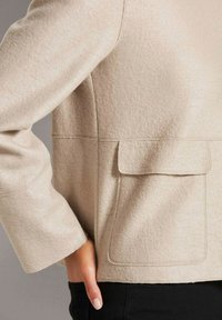 Beige wollenblend jasje met een gestructureerde afwerking, een ontspannen pasvorm en een patchzak, gecombineerd met zwarte broeken.