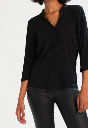 Blusa nera a maniche lunghe con scollatura a V e dettagli con bottoni, realizzata in tessuto liscio; indossata con leggins neri lucidi.