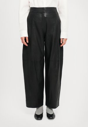 Pantalons larges en cuir noir à taille haute, avec des détails cousus et une texture lisse, associés à des chaussettes grises et des chaussures noires.