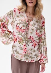 Bluse aus leichtem Stoff mit floralem Muster, verfügt über einen V-Ausschnitt mit Schnürdetails, lange Puffärmel und einen geschwungenen Saum. Die Muster zeigen rosa und grüne Blumen.