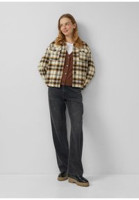 Kurtka w kratę w kolorach kremowym, brązowym i czarnym z zapięciem na guziki, zestawiona z brązowym kardiganem i czarnymi jeansami, do tego masywne czarne buty.