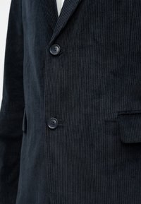 Blazer em veludo cotelê azul escuro com superfície ligeiramente texturizada, dois botões e lapelas com entalhe. Inclui bolsos frontais.