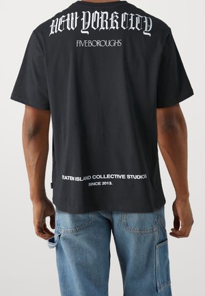 Persoon draagt een zwart t-shirt met witte tekst "New York City Five Boroughs" en "Staten Island Collective Studios Since 2013" op de achterkant.