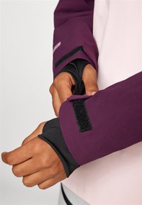 Dve roke prilagajajo manšete vijoličnega jopiča z notranjimi rokavi v črni barvi in velcro zapiralom, kar prikazuje plasten dizajn tkanine.