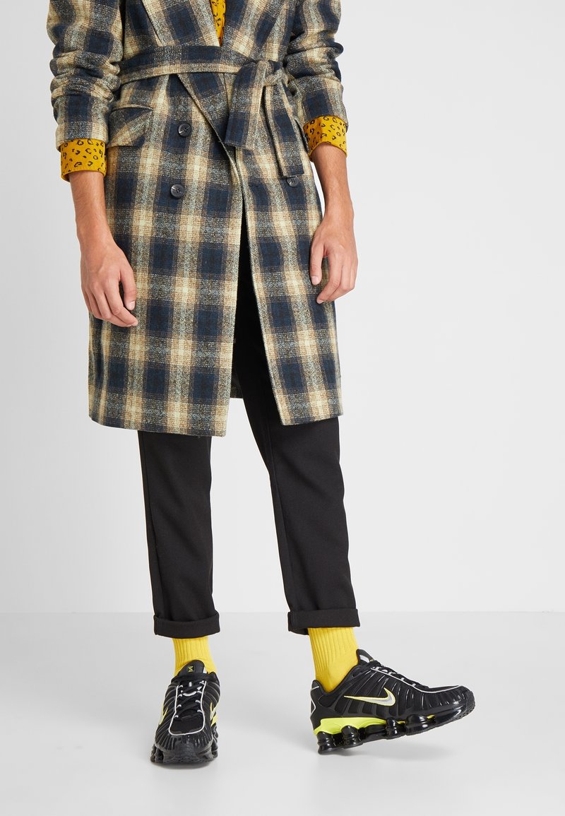 Personne portant un manteau à carreaux, un pantalon noir aux ourlets retroussés, des chaussettes jaunes et des baskets noires avec des détails jaunes sur un fond uni.
