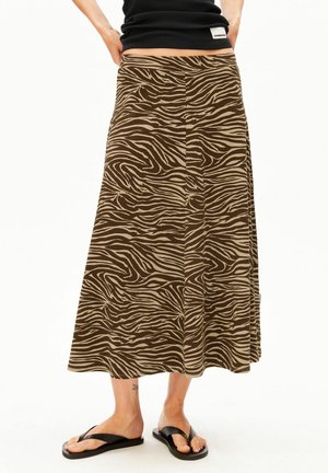 Frau trägt einen braun-beigen, zebragestreiften Midi-Rock, ein schwarzes Top und schwarze Flip-Flops, steht mit den Händen in der Hüfte.