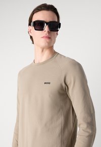 Bež srajca z dolgimi rokavi s teksturiranim vzorcem in črnimi logotipom "BOSS". Model nosi prevelike črne sončna očala. Preprost, moderen dizajn.