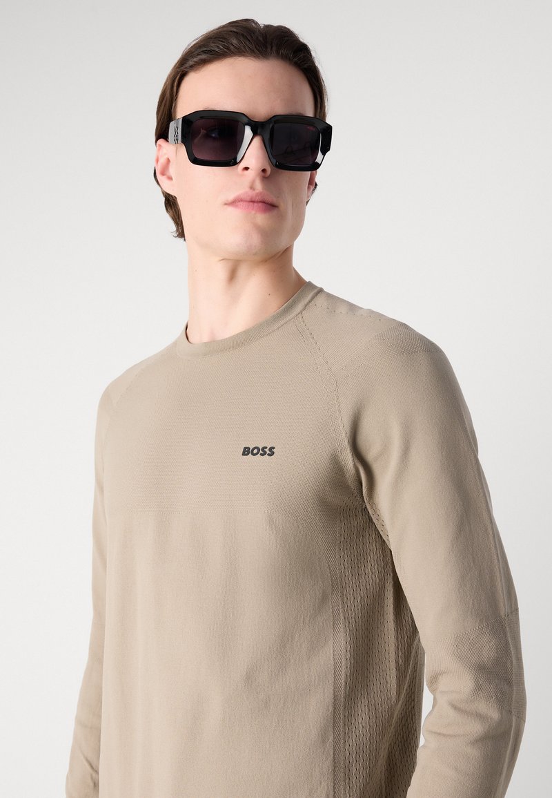 Camisa de manga larga beige con un patrón texturizado y el logo negro de "BOSS". Modelo llevando unas gafas de sol negras de gran tamaño. Diseño sencillo y moderno.