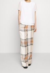 Haut blanc à manches courtes avec des manches transparentes associé à un pantalon large à carreaux dans des tons beige, marron et noir, présentant une texture lisse.