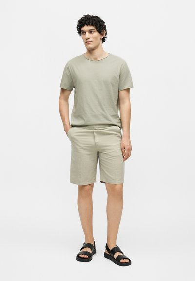 Νέος άνδρας με σγουρά μαλλιά που φοράει ανοιχτό πράσινο μπλουζάκι, αντίστοιχα σορτς και μαύρα σανδάλια, στέκεται με τα χέρια στις τσέπες.