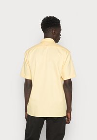 Camisa de mangas curtas em amarelo pálido, tecido texturado, corte reto, colarinho abotoado, com um simples yoke nas costas e linhas limpas.