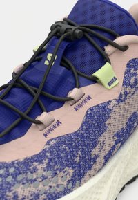 Chaussure de sport avec une tige violette texturée, des accents roses et des motifs gris, dotée de lacets noirs sécurisés et d'une boucle de fixation noire.
