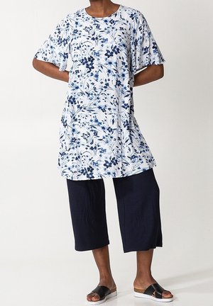 Persoon die een witte tuniek draagt met blauwe bloemenpatronen, marineblauwe wijde cropped broek en zwarte slippers, staand met de handen achter de rug.