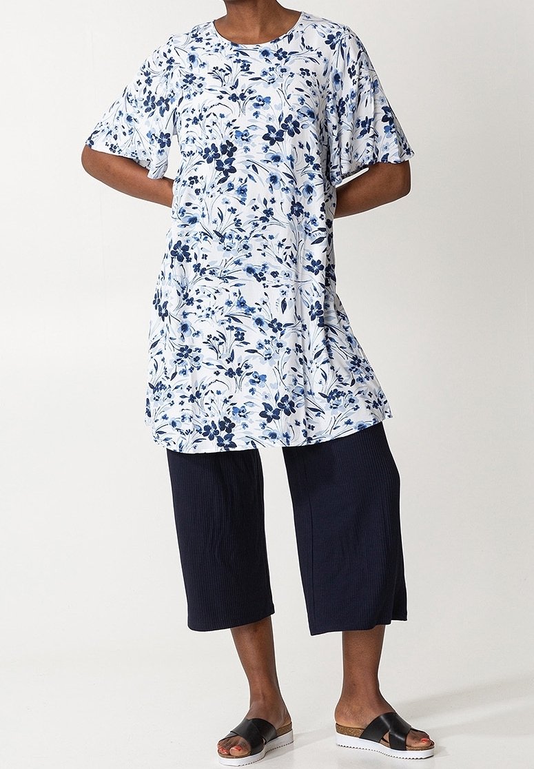 Personne portant une tunique blanche avec des motifs floraux bleus, un pantalon large court bleu marine et des sandales noires, debout les mains dans le dos.