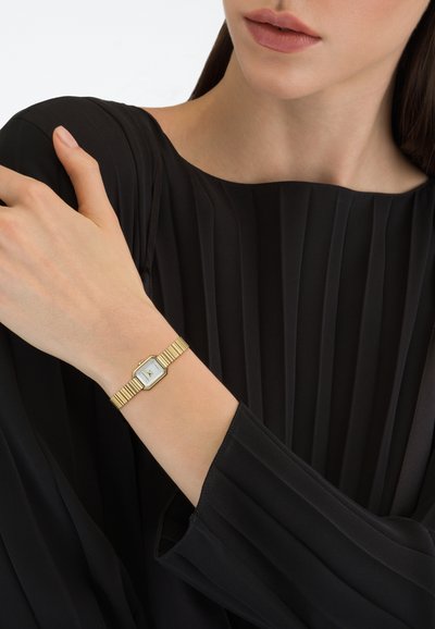 Aranyszínű hatszögletű karóra fehér számlappal és fekete mutatókkal, texturált fém szíjjal, világos bőrszínű csuklón, fekete pliszírozott szövet mellett viselve.