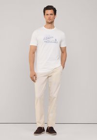Camiseta blanca de manga corta con gráficos de texto en azul, combinada con pantalones beige claros y zapatos oscuros de tela. Fondo neutro.