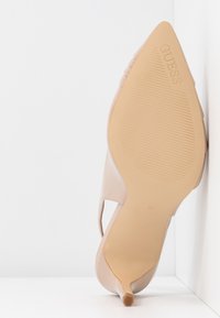 Chaussure à talon haut beige à bout pointu montrant la semelle texturée avec le logo "GUESS" et la taille 37 sur une surface et un mur blancs.