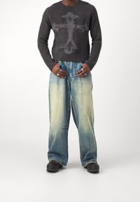 Maglione in lana nera, strutturato, con un disegno centrale di una croce grigio chiaro; jeans svasati blu sbiaditi; scarpe nere con suole spesse; anelli d'argento.