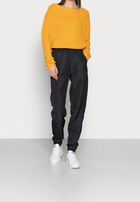 Žlutý pletený svetr s širokým výstřihem a dlouhými rukávy, v kombinaci s černými saténovými tepláky a bílými teniskami, stojící na světle šedé podlaze.