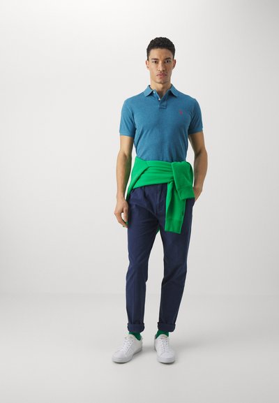 Polo de color aguamarina con mangas cortas y un logo rojo, combinado con pantalones azul marino y zapatillas blancas, acentuado por un suéter verde atado a la cintura.